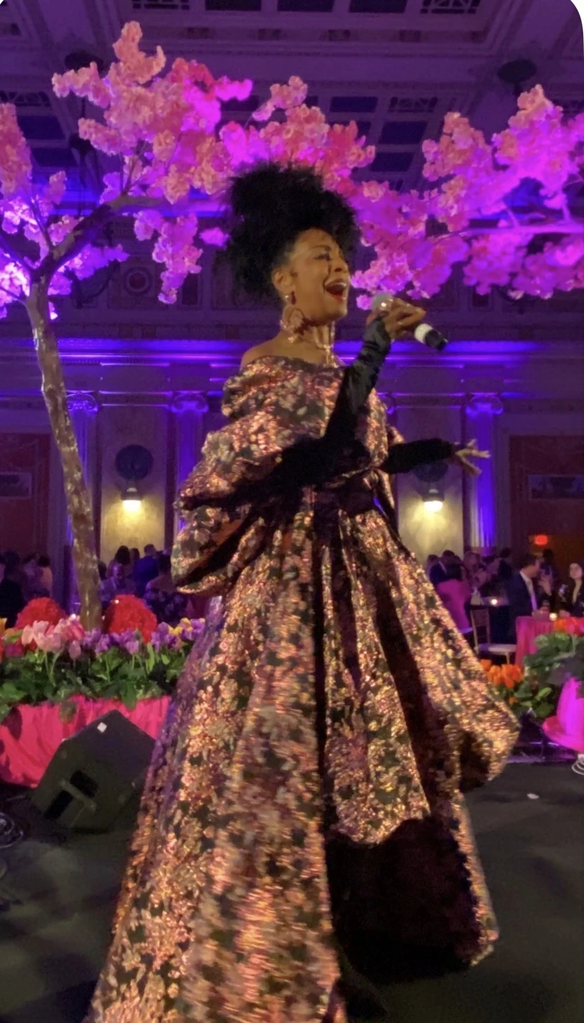 Yvette Spears singing beneath pink blossoms at a gala, wearing a richly patterned gown